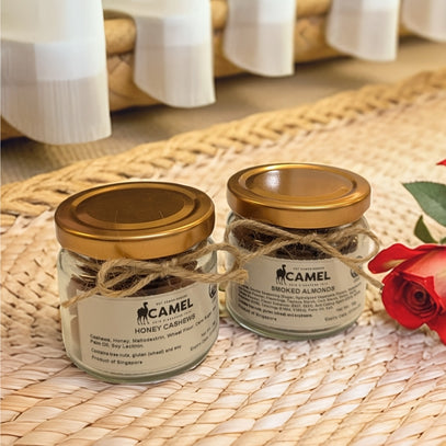 Two jars of Camel Nuts honey cashews and smoked almonds on a woven mat with a red rose.
