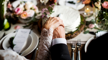 Bride and groom holding each other's hands with a wedding cake and floral arrangements in the background. Celebrate your wedding with Camel Nuts premium nuts in Singapore