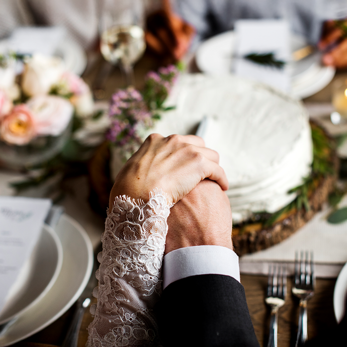 Two hands holding each other with a wedding cake and floral arrangements in the background. Celebrate your wedding with Camel Nuts premium nuts in Singapore