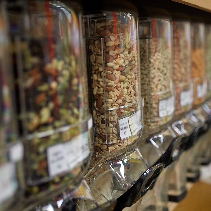Row of dispenser jars with various types of nuts and seeds on a shelf. At Camel Nuts we have the best variety of nuts for your business. Order in bulk.