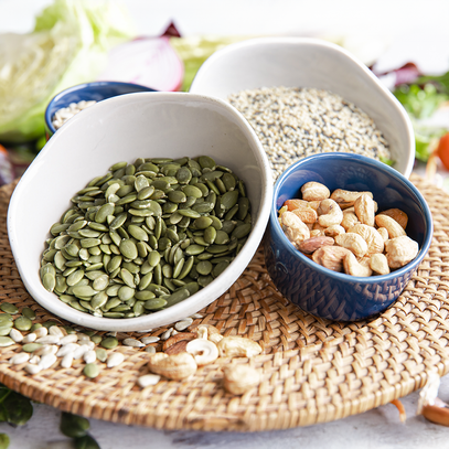 Bowls of pumpkin seeds and cashews on a woven placemat with blurred seeds and vegetables in the background. Find your baking needs and ingredients at Camel Nuts