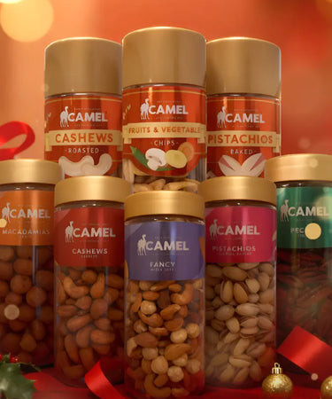 Camel brand food products including cashews, fruits & vegetables, pistachios, and fancy mix on a red background with festive decorations.