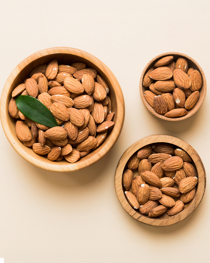 Camel Nuts Singapore almonds in wooden bowls – Three wooden bowls filled with almonds, with a green leaf accent.