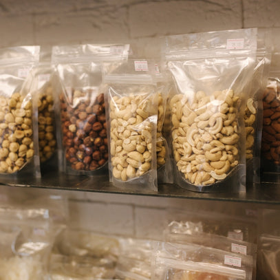 Assorted nuts in clear plastic bags on a shelf with a stone wall background. Camel Nuts Singapore