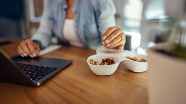 Person sitting at a desk with a laptop, holding a bowl of premium hazelnuts and other nuts
