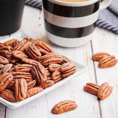 Pecans in a white dish with a black and white striped mug on a wooden surface. Camel Nuts Singapore