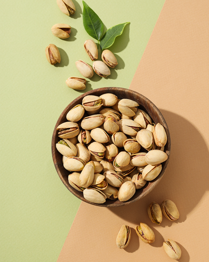 Camel Nuts Singapore pistachios in bowl with leaves – Whole pistachios in a wooden bowl with green leaves on pastel background.